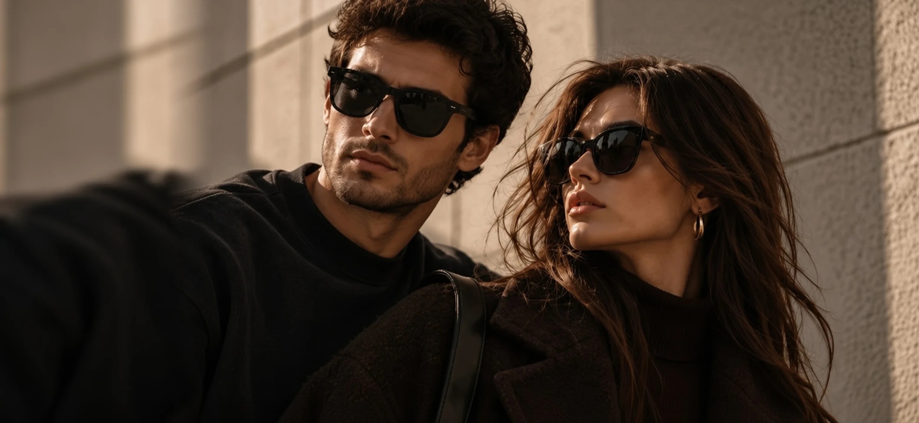 A couple in sunglasses and dark knitwear against a sunlit stone wall.